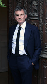 Cyril prend la pose davant la porte d'entrée d'une maison à Trouville et porte le costume droit Barthélémy en flanelle de laine coloris bleu marine - collection automne-hiver 2024 - de la Maison Gabriel Paris