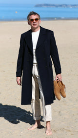 Cyril sur la plage de Trouville tient la paire de Richelieu Weston en veau velours couleur noisette, et porte le manteau polo-coat Bart en laine et cachemire coloris bleu nuit de la collection Automne-Hiver 2024 de la  Maison Gabriel Paris