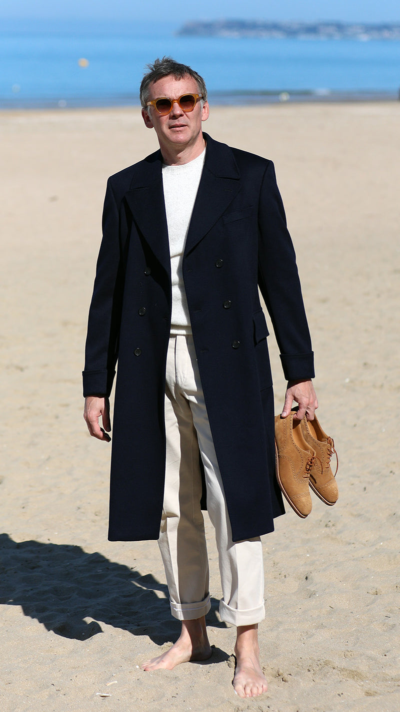 Cyril sur la plage de Trouville tient la paire de Richelieu Weston en veau velours couleur noisette, et porte le manteau polo-coat Bart en laine et cachemire coloris bleu nuit de la collection Automne-Hiver 2024 de la  Maison Gabriel Paris