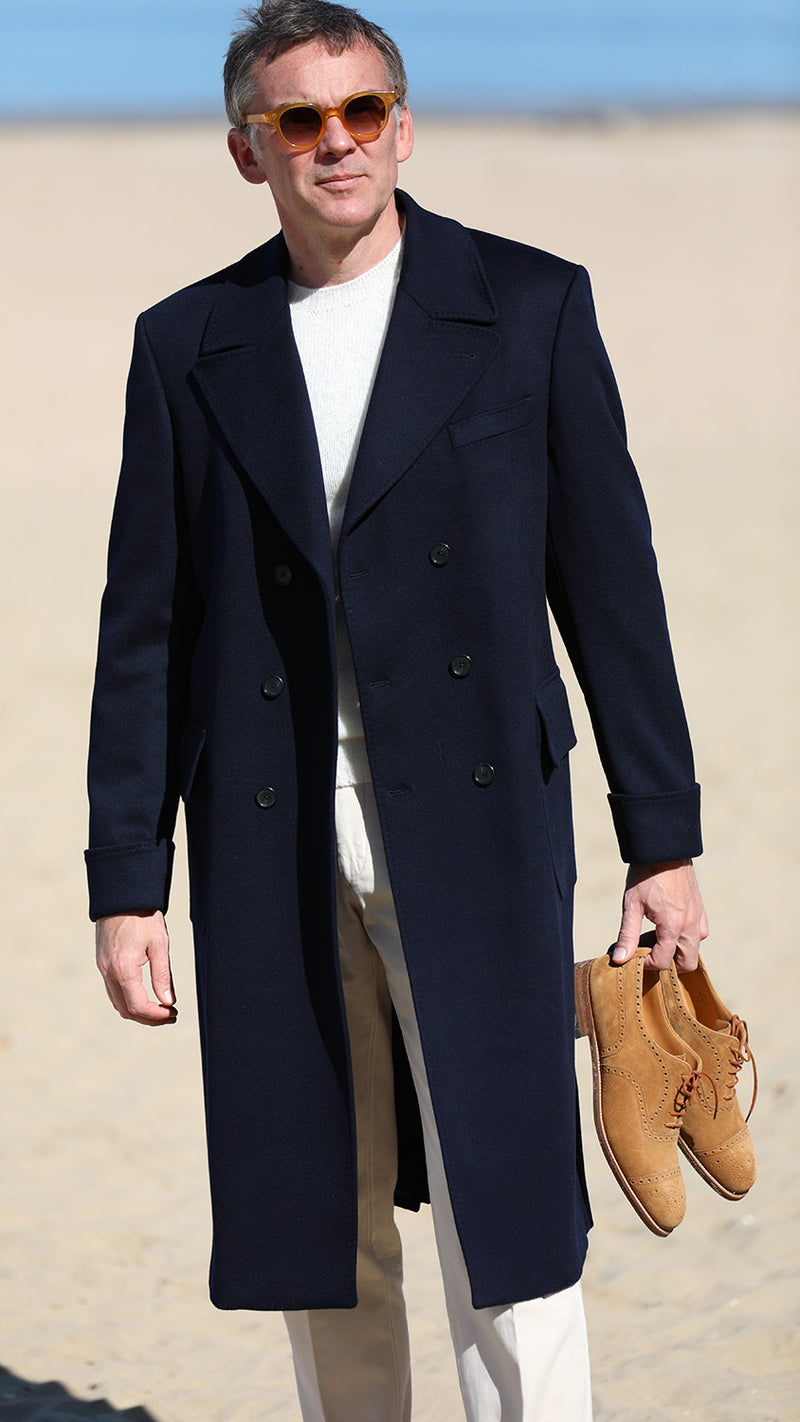 Cyril sur la plage de Trouville tient la paire de Richelieu Weston en veau velours couleur noisette, et porte le manteau polo-coat Bart en laine et cachemire coloris bleu nuit de la collection Automne-Hiver 2024 de la  Maison Gabriel Paris