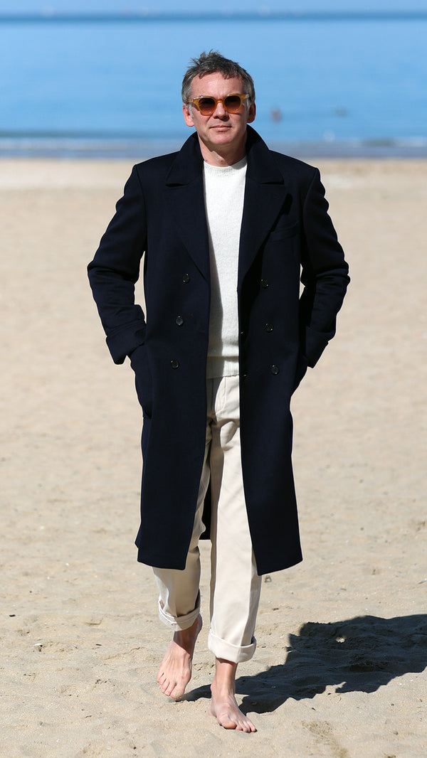 Cyril marche dans le sable sur la plage de Trouville, et porte le manteau polo-coat Bart en laine et cachemire coloris bleu nuit de la collection Automne-Hiver 2024 de la  Maison Gabriel Paris